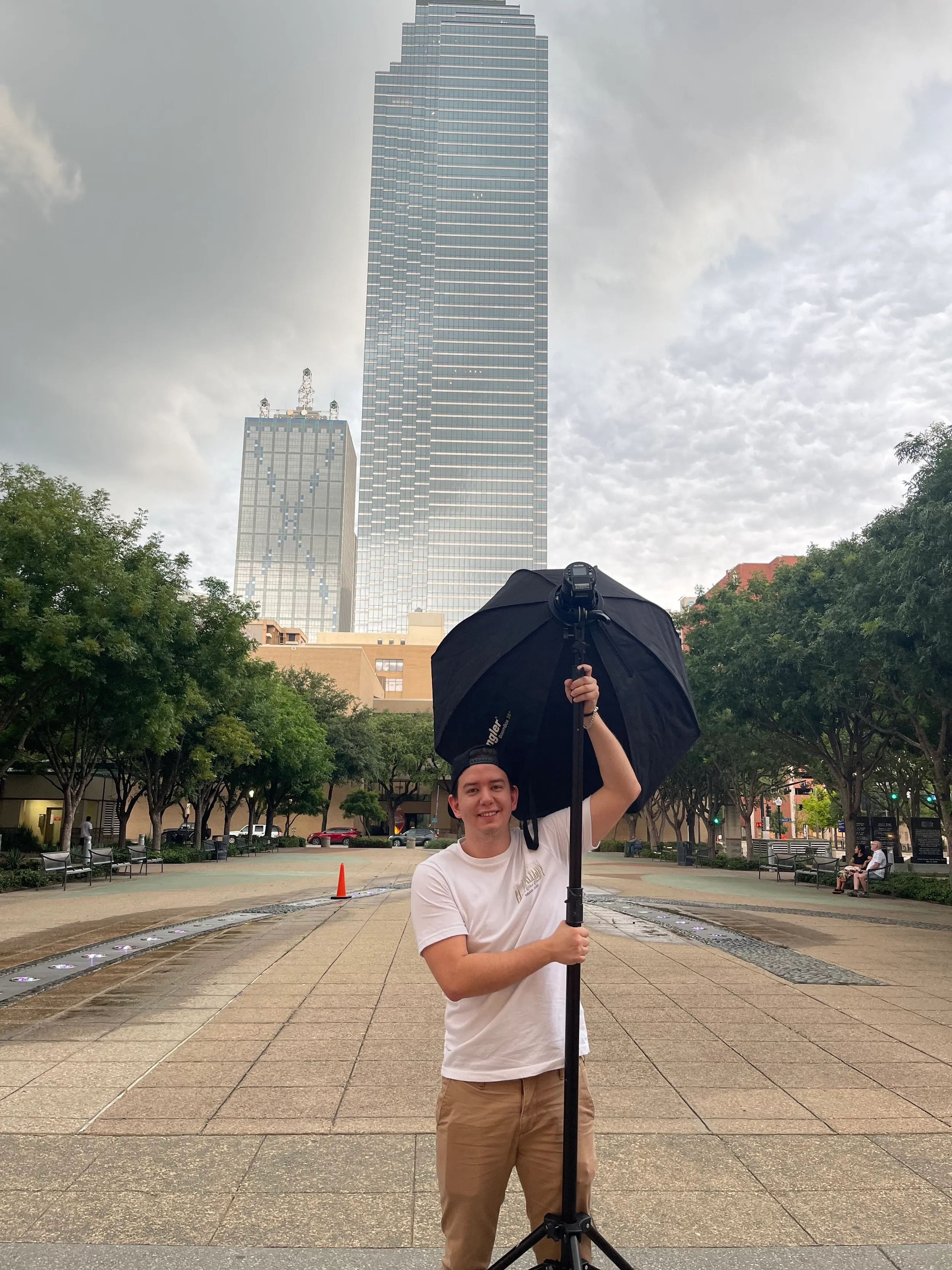 A professional photographer holding a large black umbrella and a camera mounted on a tripod at an outdoor park with tall skyscrapers in the background.