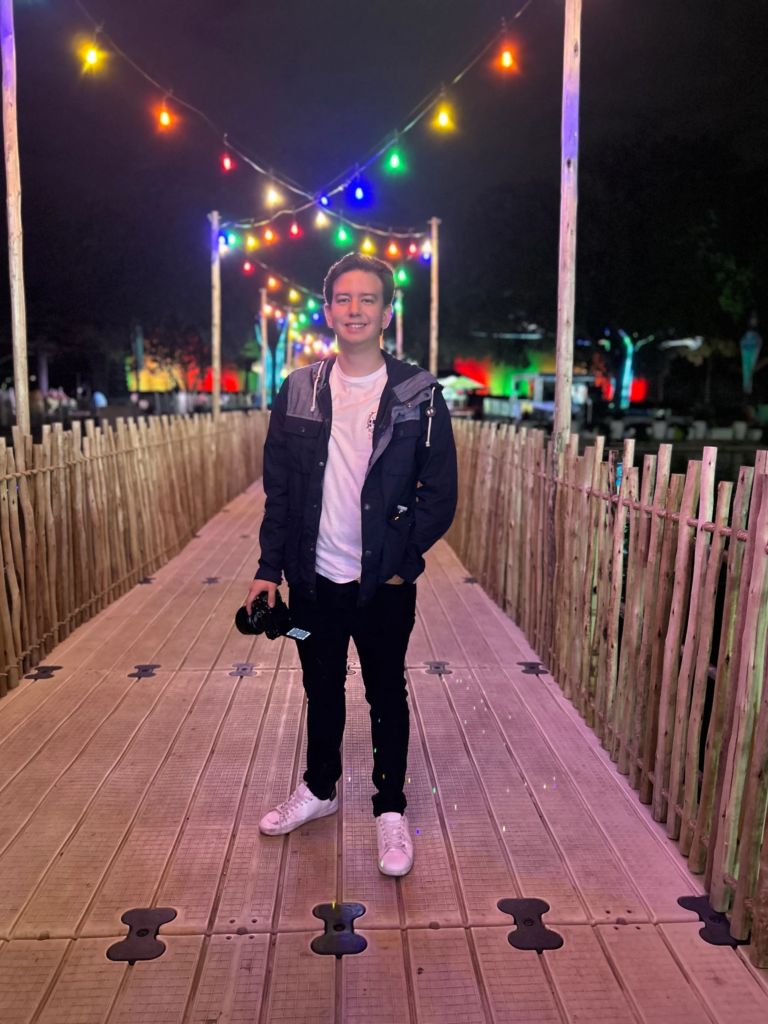 A professional young photographer standing on a wooden pathway illuminated with colorful string lights at night, holding a camera and smiling.