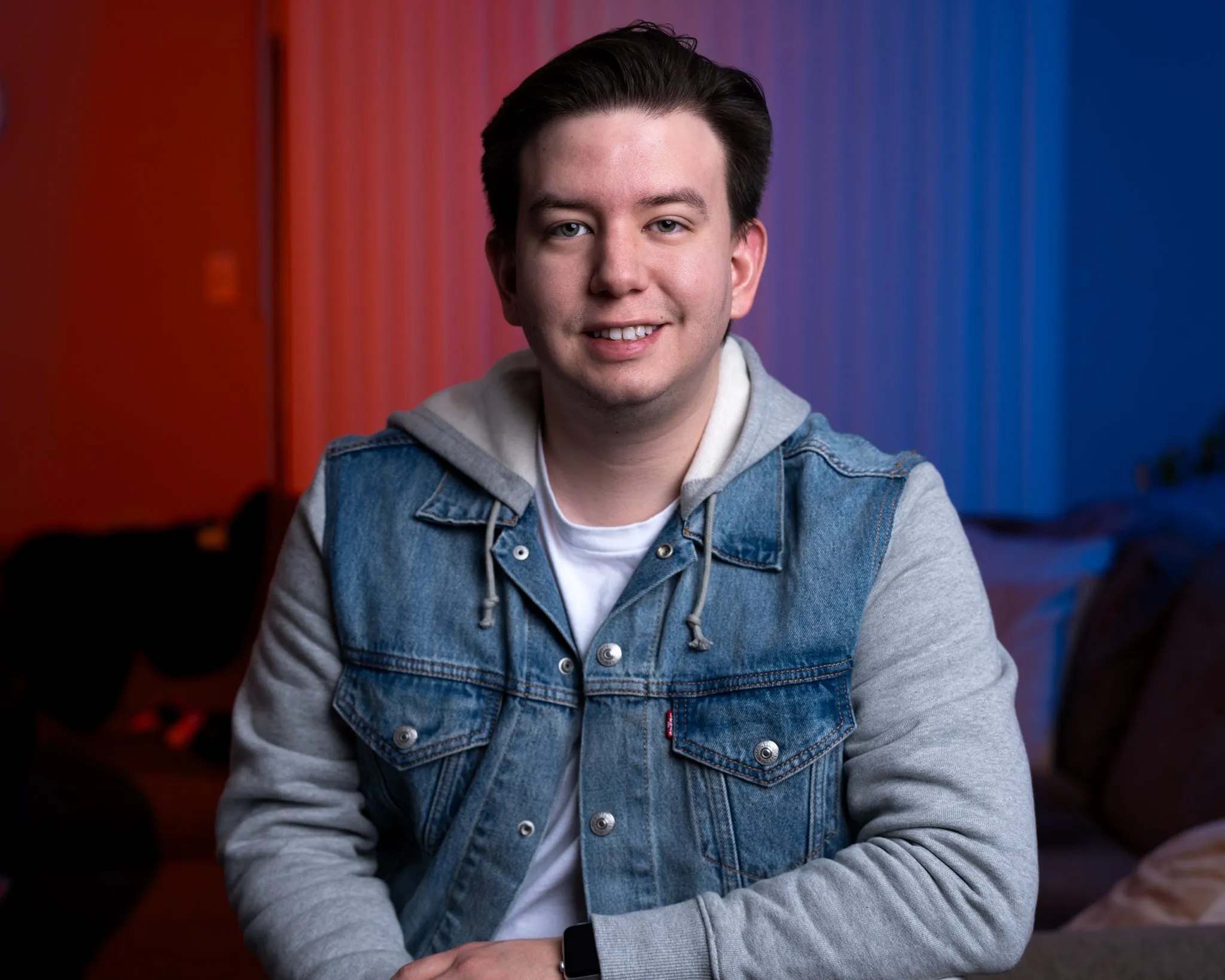 Young professional quinceañera photographer Rony Lanza sitting indoors, smiling, wearing a denim and gray hooded sweatshirt, with red and blue lighting in the background.