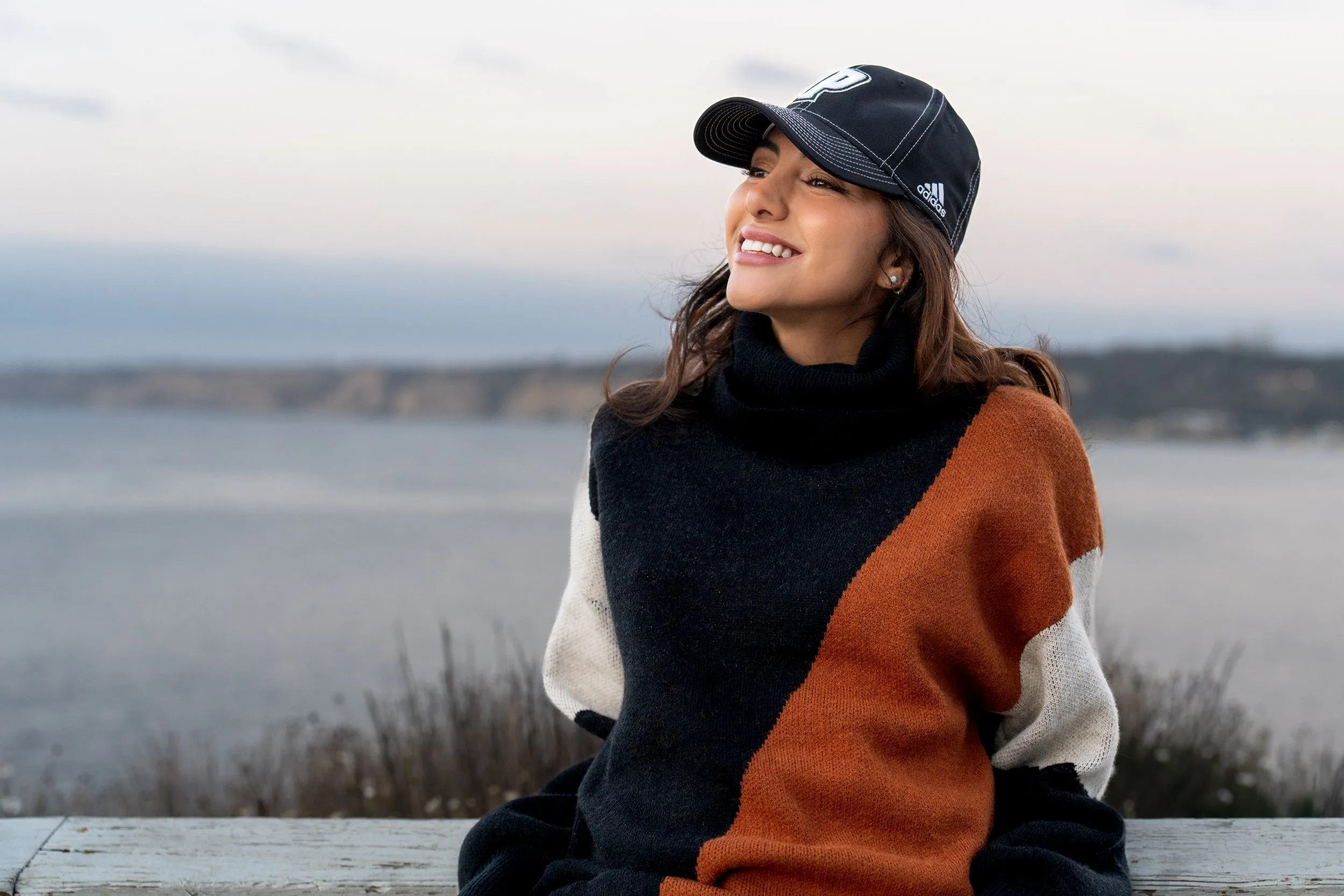 A woman with brown hair wearing a black baseball cap and a color-blocked sweater, sitting outdoors near water during a portrait session
