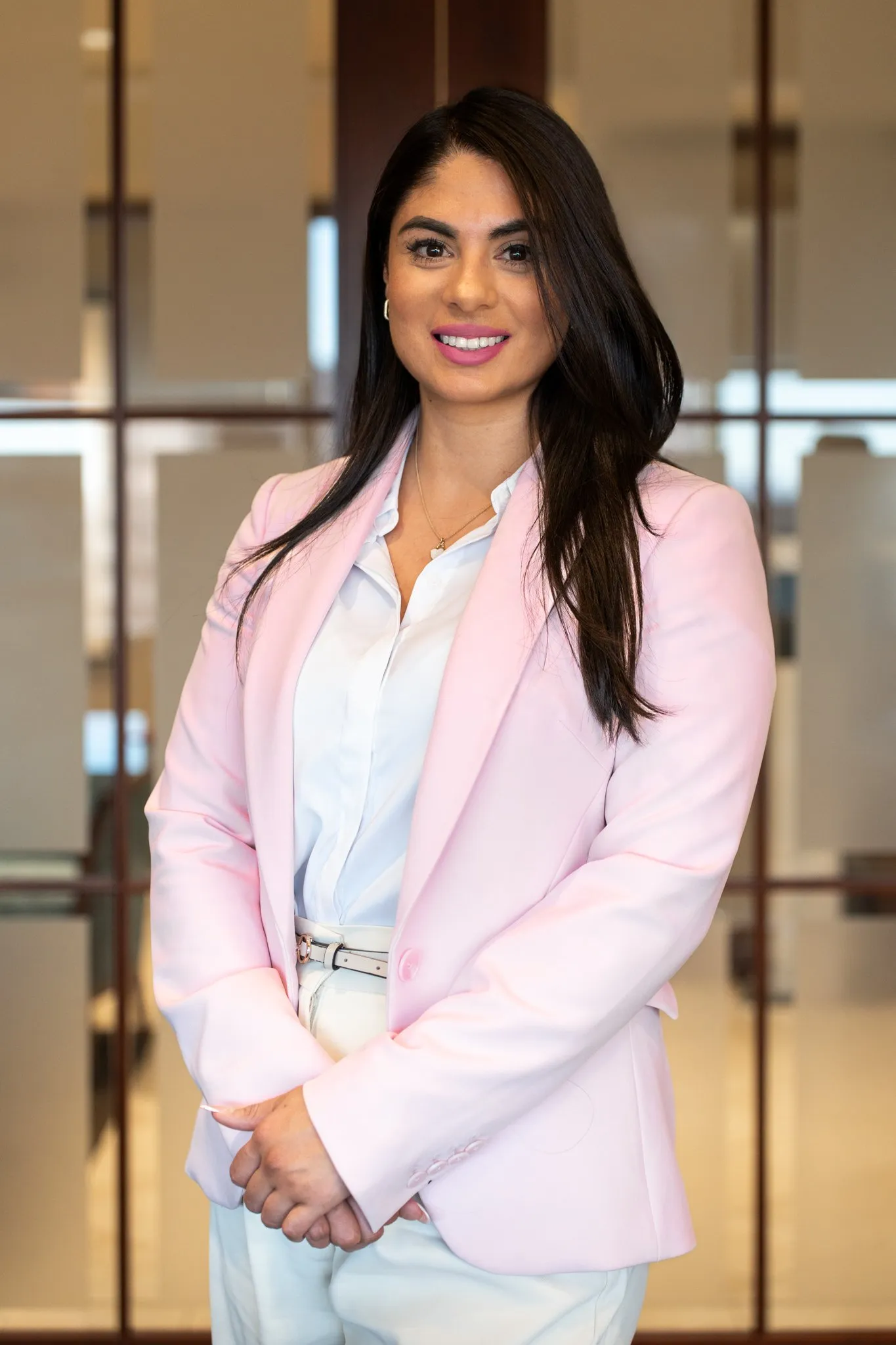 A woman with long dark hair, wearing a light pink blazer, white shirt, and white pants, standing indoors against a background with glass panels and a modern office setting posing for her professional headshots.