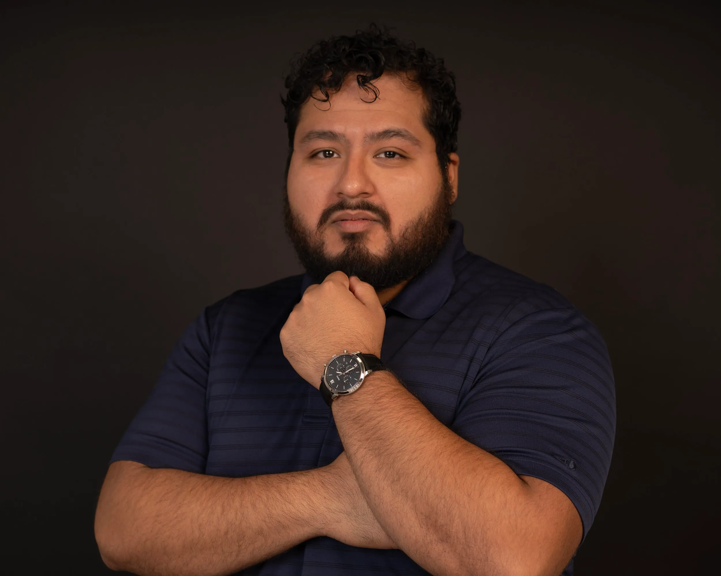 A man with curly hair and a beard wearing a navy blue polo shirt and a black wristwatch, standing against a dark background, with one hand resting on his chin and the other crossed over his chest.
