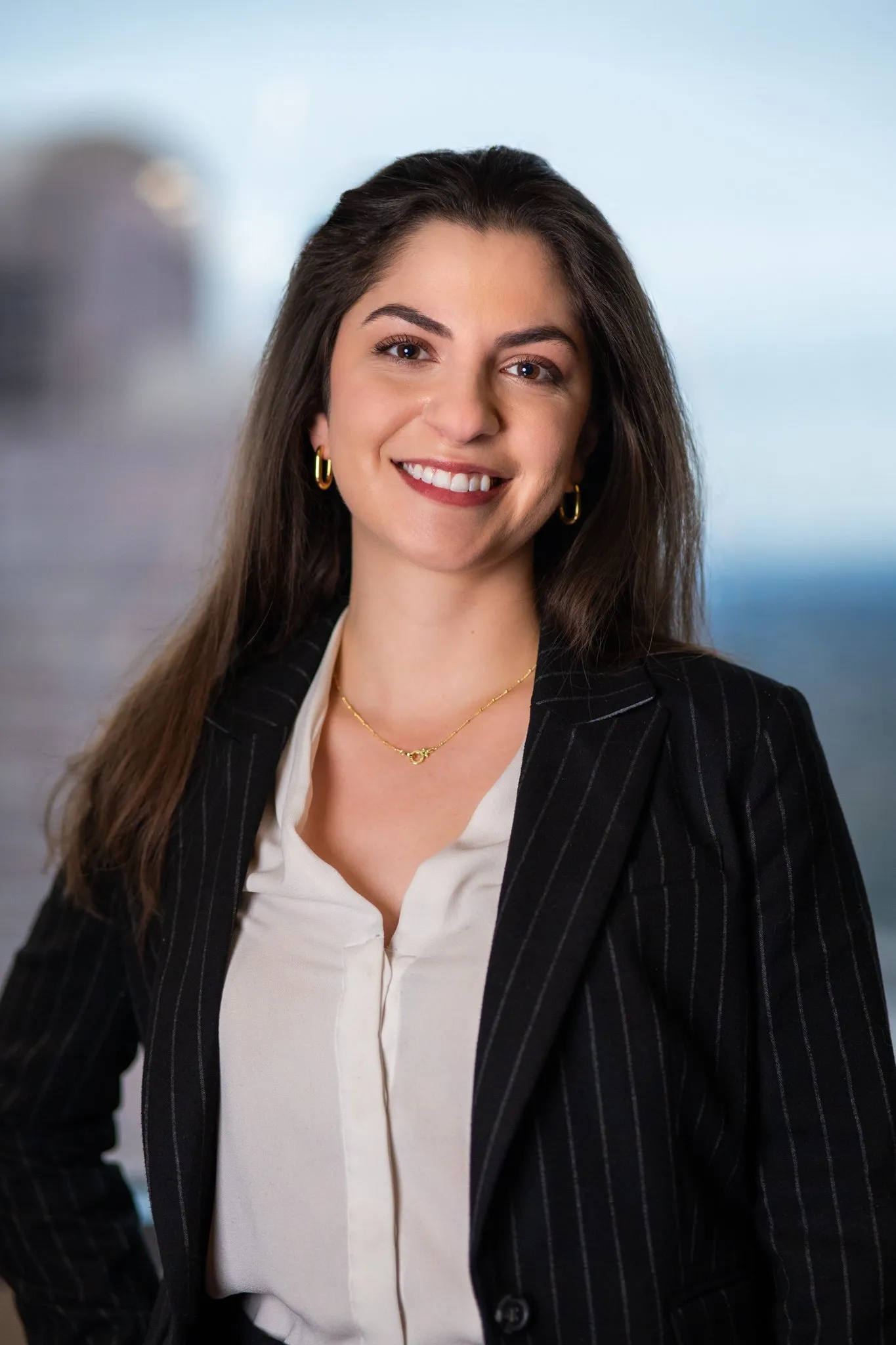 A woman with long dark hair, smiling, wearing a black pinstripe blazer, white blouse, gold jewelry, standing against a blurred background that appears to be a cityscape or waterfront posing for her professional headshots.