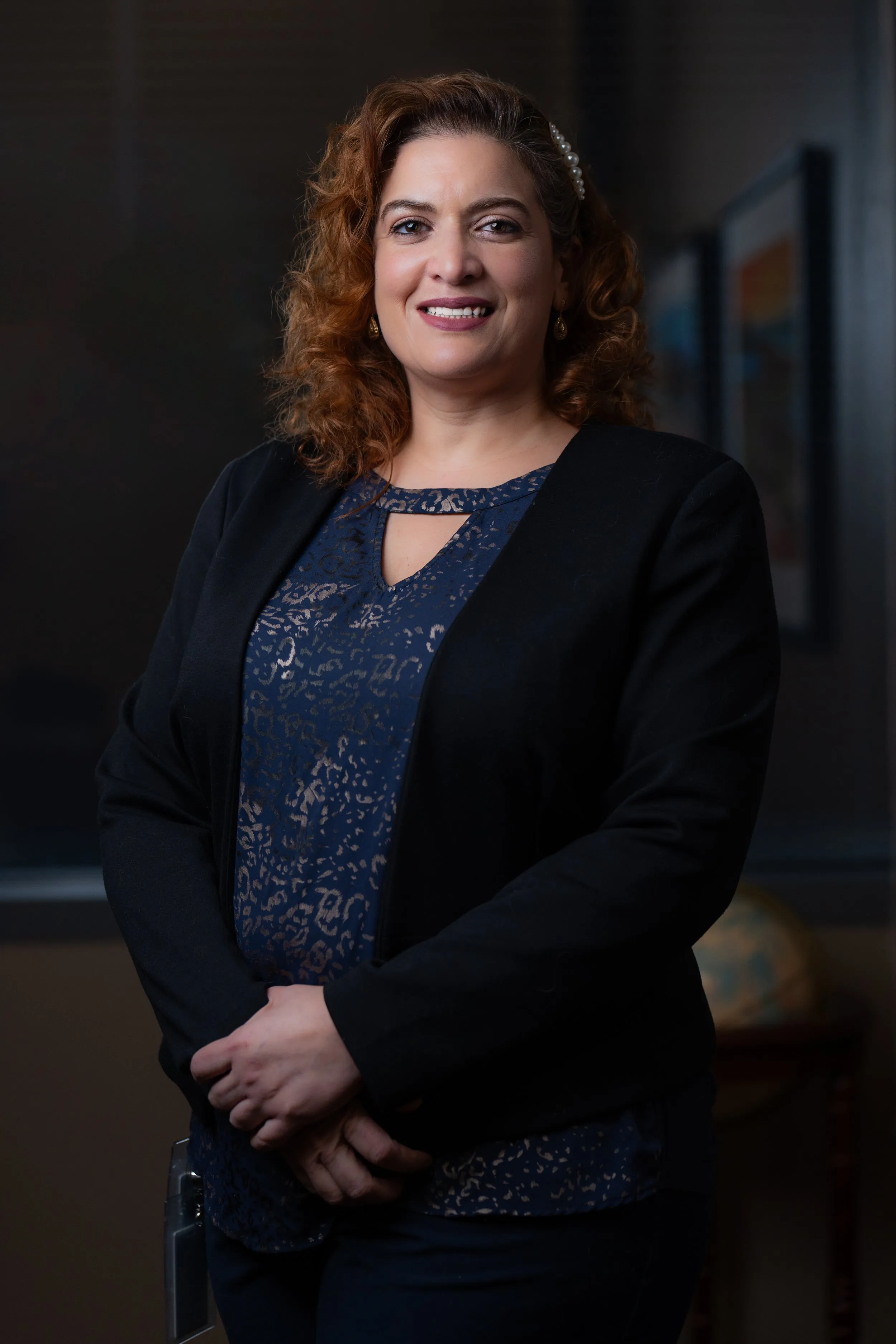 A woman with curly red hair wearing a navy blue and black outfit standing in an office or conference room posing for her professional headshots.