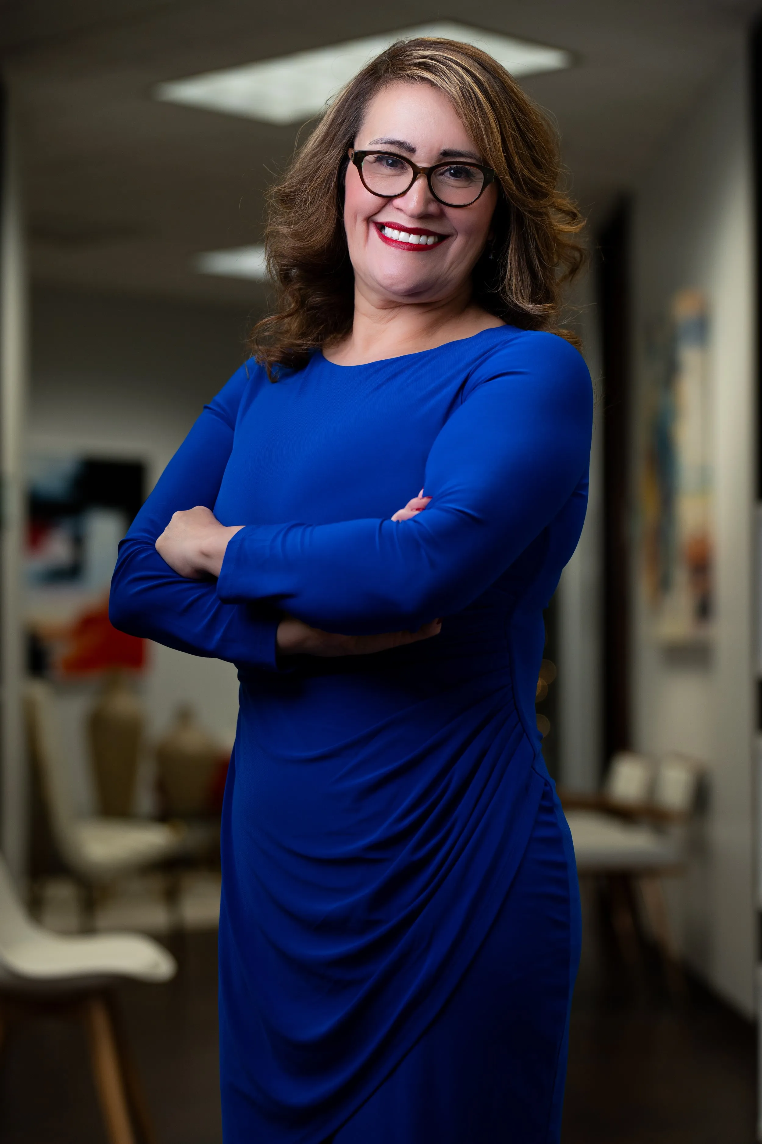 A smiling woman with glasses in a blue dress standing with arms crossed in an office setting posing for her professional corporate headshots.